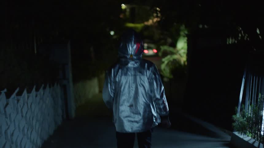 Lights flash and strobe as a person wearing a silver jacket with hood up walks down a sloped driveway toward the street at night