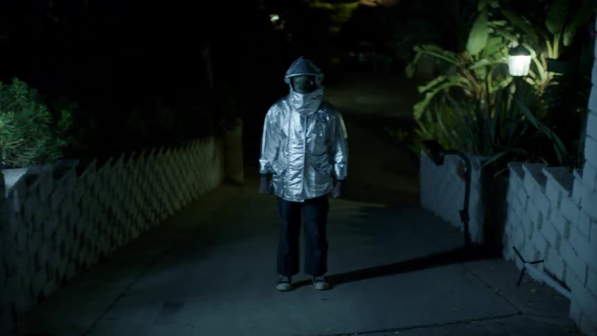 Person wearing hooded jacket and full face mask is revealed by flickering lights standing in driveway between fences at night