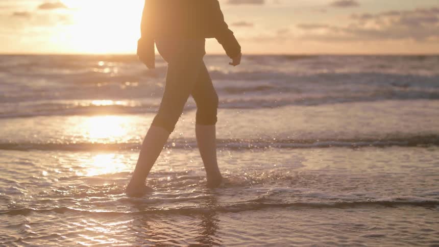 A woman walking at the water's edge on a beach at sunset