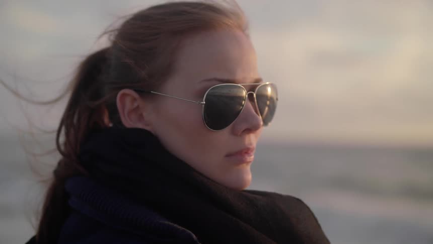 A woman wearing sunglasses is standing by the water at sunset