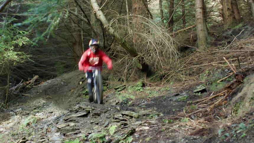 Person riding bike races around corner in woods taking jump