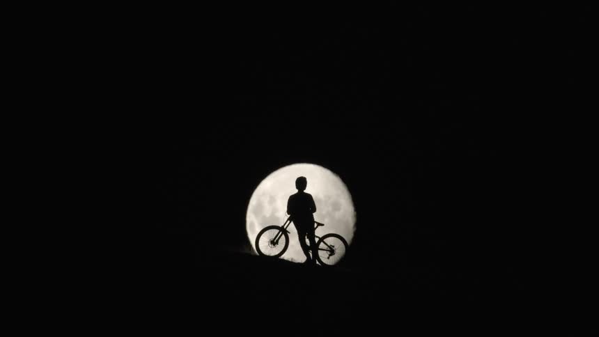 Young person leaning against bike silhouetted by moon