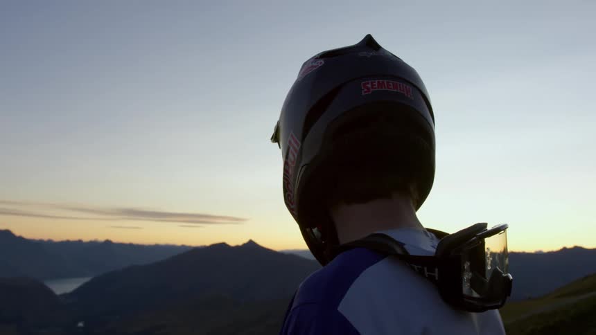 Man wearing crash helmet looks at valley from a mountain at sunset
