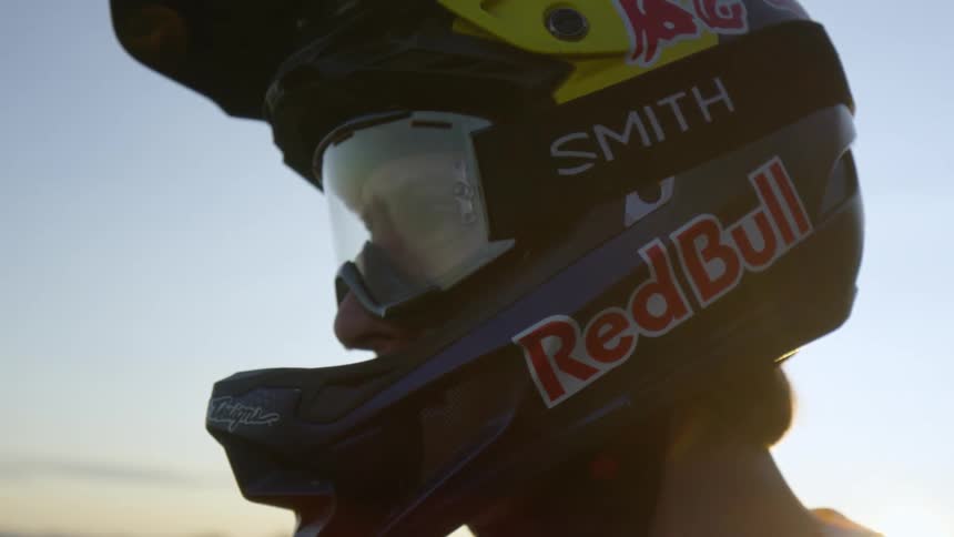 A competitive motorcyclist stands in a desert near a mountain range wearing a helmet adorned with sponsors