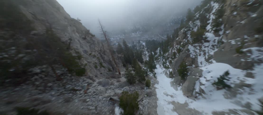 A narrow fog shrouded valley slope between mountains leading down to a much wider valley