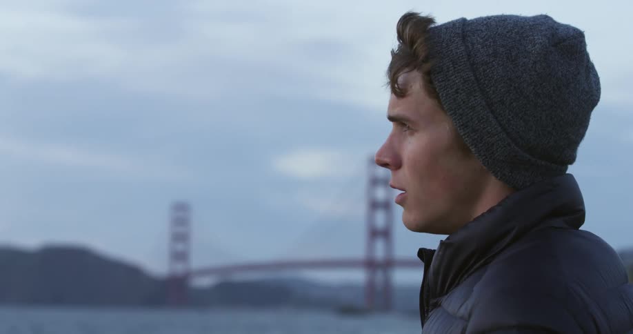 A troubled young man stands alone on a shore with the golden gate bridge in the background