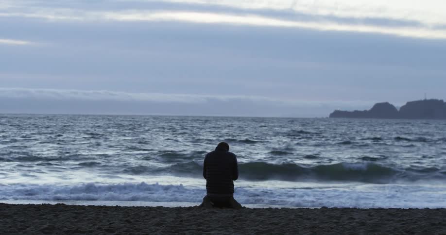 Stressed man on his knees on the beach is silhouetted against the wavy sea at dusk