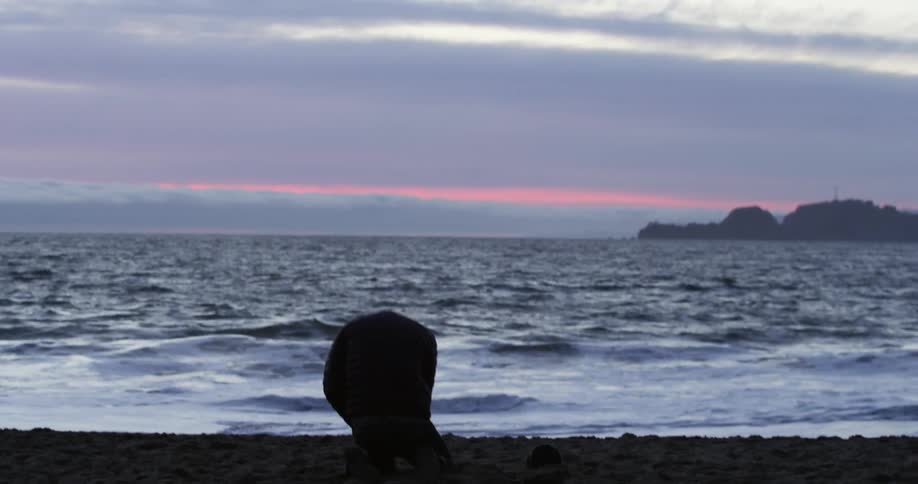 A man walking on the shore towards the water drops on his knees and hits the sand and cries