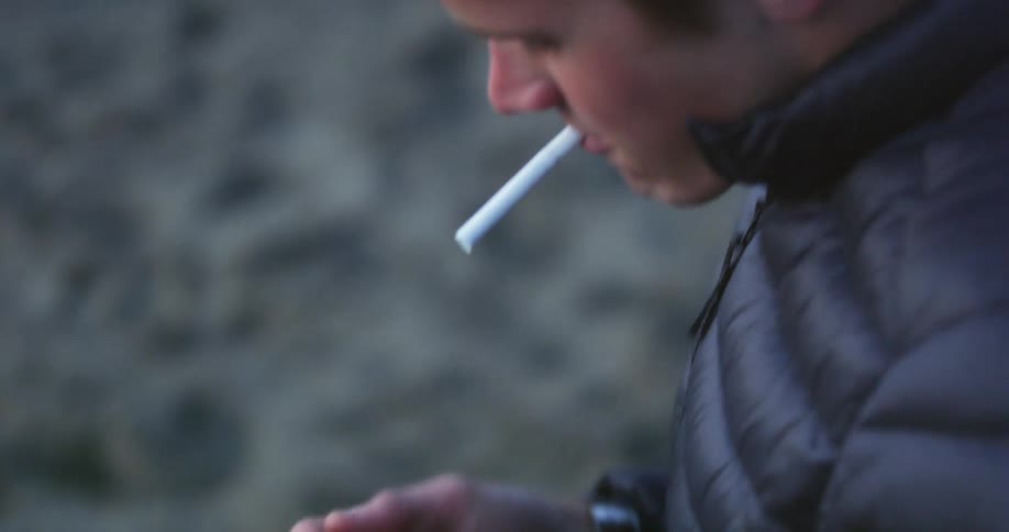 Young man tries to light a cigarette with a lighter while standing in the sand of a beach