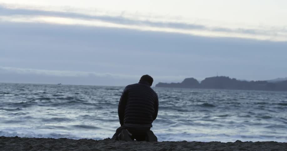Man kneels on the sand of a beach at sunset