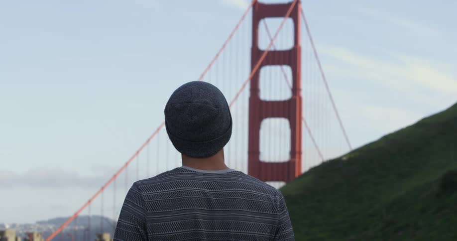 A man stands on a hilltop looking towards a large city bridge
