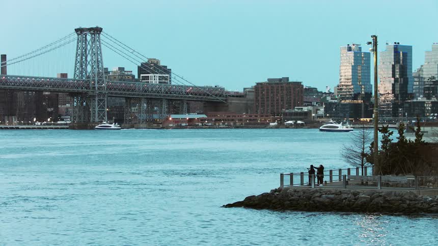 Two people stand on the shore and point as boats pass on the water and cars drive across a bridge in front of the city skyline