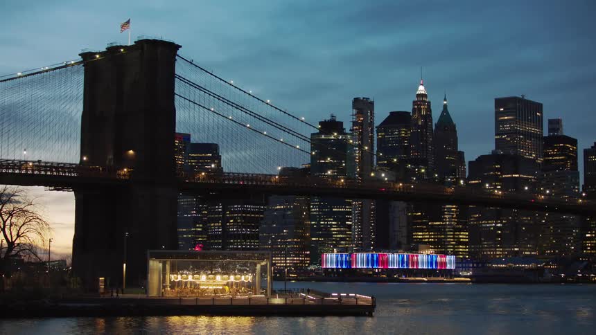 Suspension bridge that is supported by strong steel ropes hung from a tower with illuminated tall buildings in the background at dusk