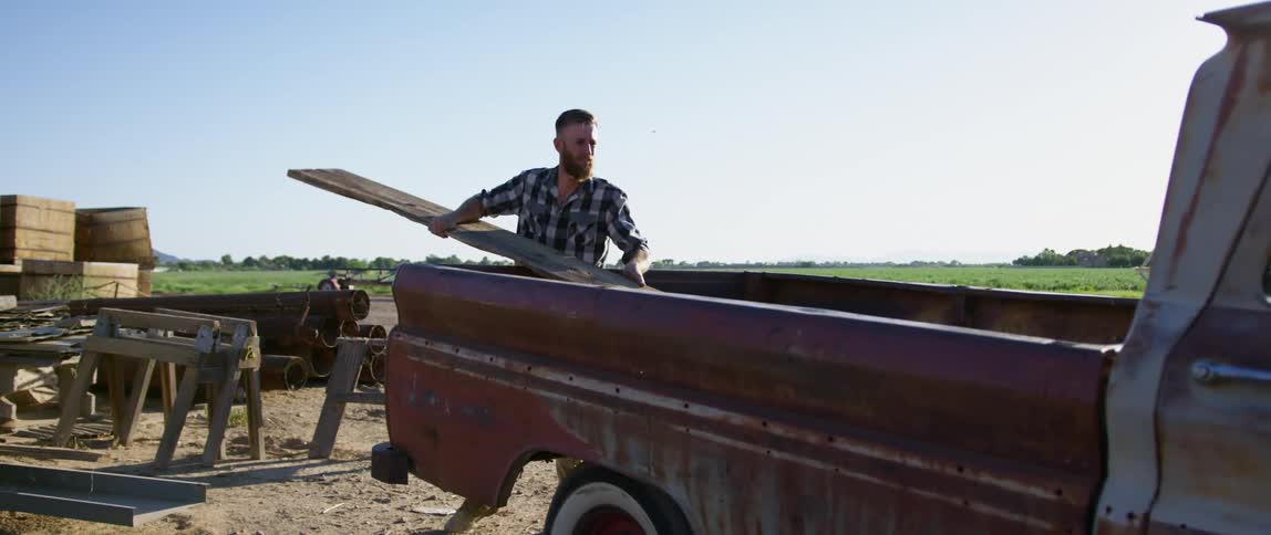 A man lifting a long wooden board to place it in the bed of a old pickup truck