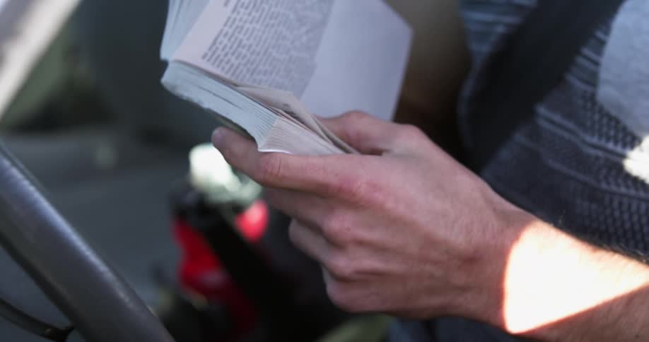 Male sitting in a stopped vehicle with his seat belt on turns the pages in a book he is reading as the sunlight shines onto him
