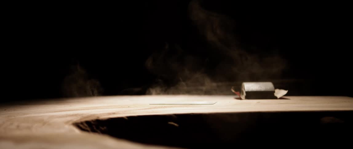 A hand places a piece of metal on a curved wooden plank next to a roll of sandpaper causing dust to rise