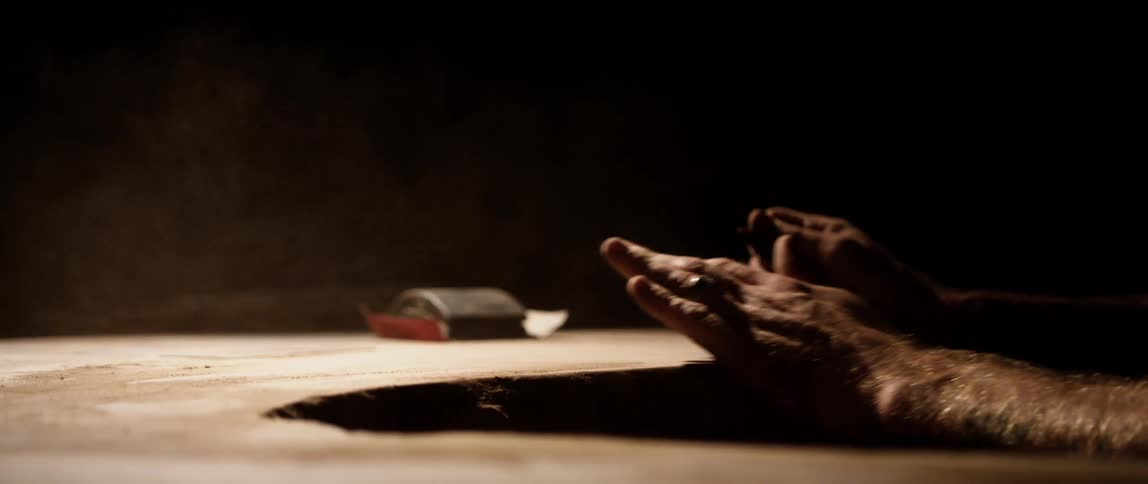 Carpenter blows sawdust from wood surface on a dimly lit room