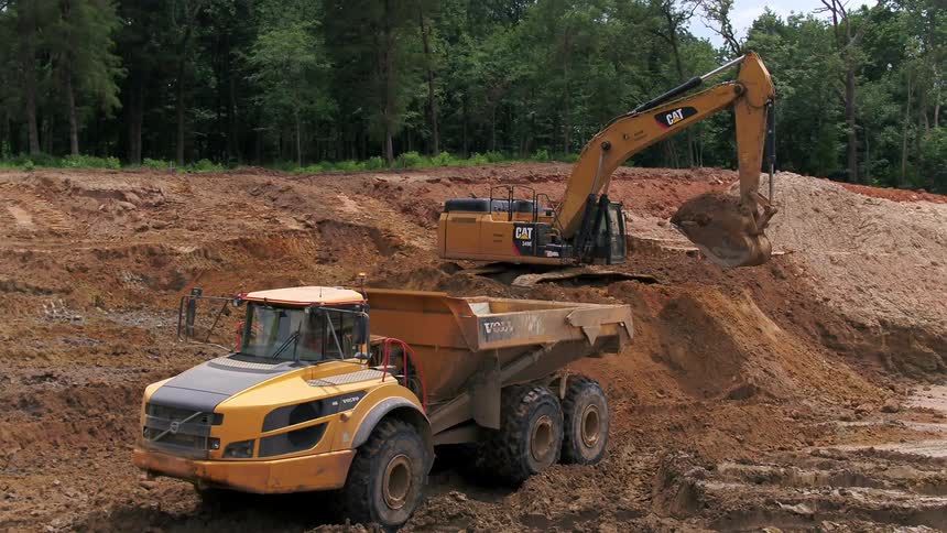 A dump truck backs up to a tractor in front of a forest