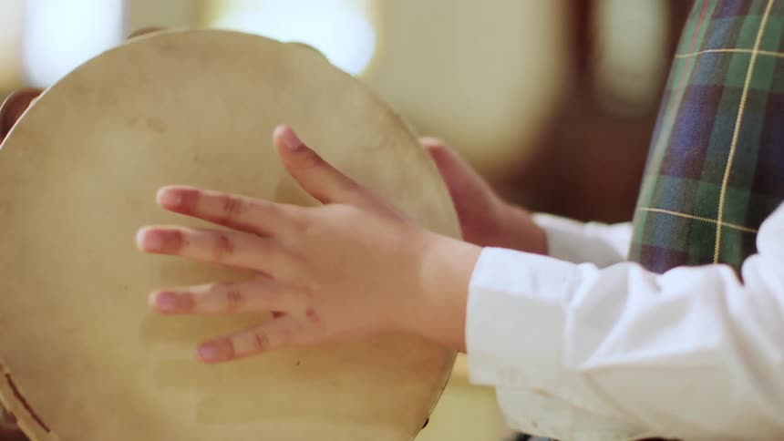A young boy in a plaid school uniform repeatedly hits a cymbal with his hand in a classroom