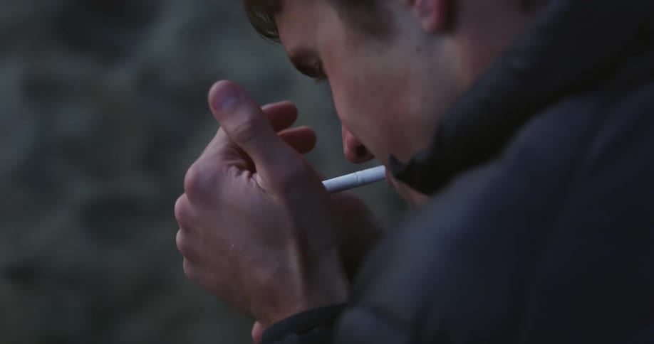 A man with a blue coat and shaky hands tries to light up a cigarette