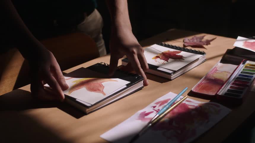 A teacher places a book and paper down on a desk next to water color paints brushes and leaves