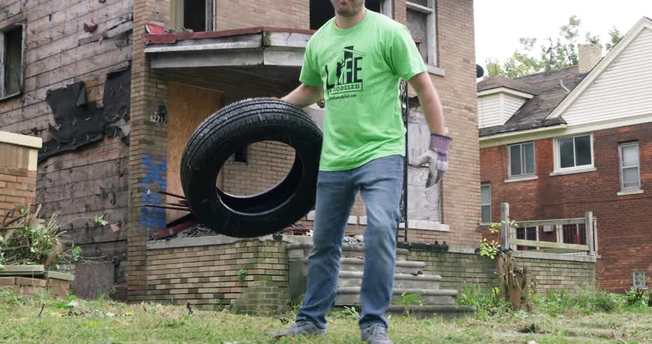 A man wearing work gloves removes a tire from the front yard of an abandoned home