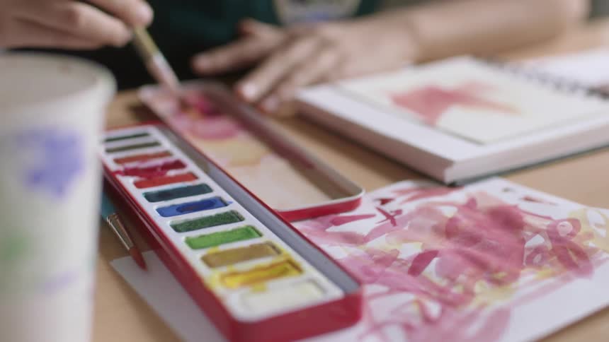 A person swirls a paintbrush into water color paint sitting at a table