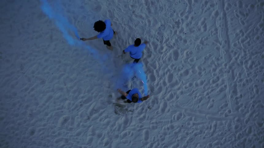Three teens run and roll in the sand while spraying blue dye in the air