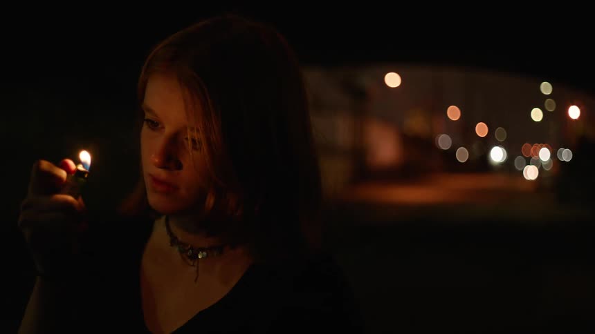 A young white woman sits in a vehicle and stares at a lighter flame