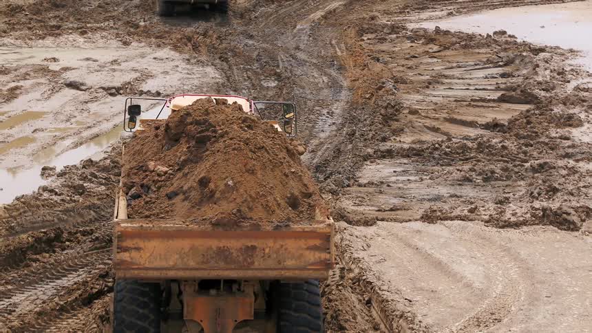 A loaded dump truck full of dirt travels through tracked mud toward another truck