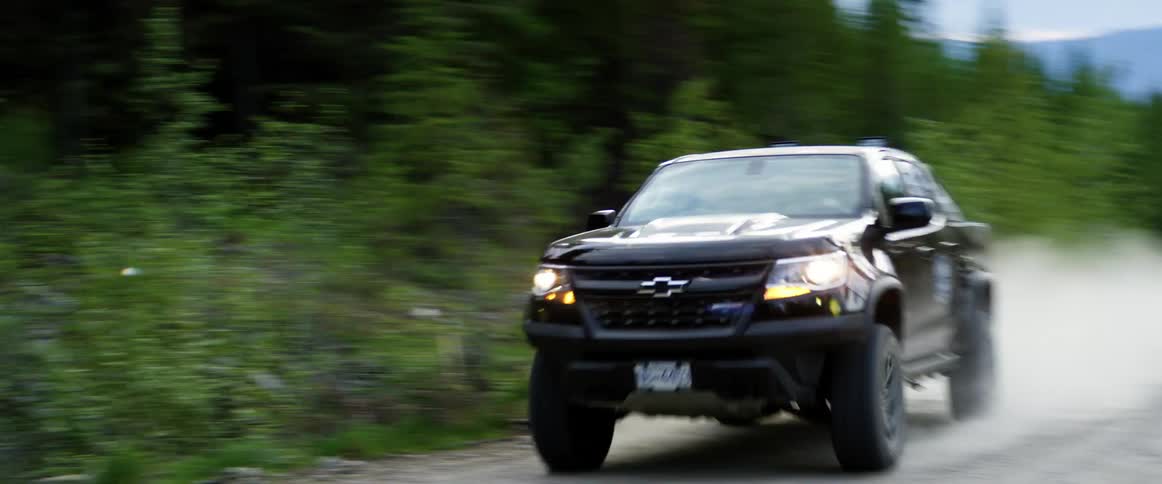 Car races down dirt road through conifer forest with mountain on horizon