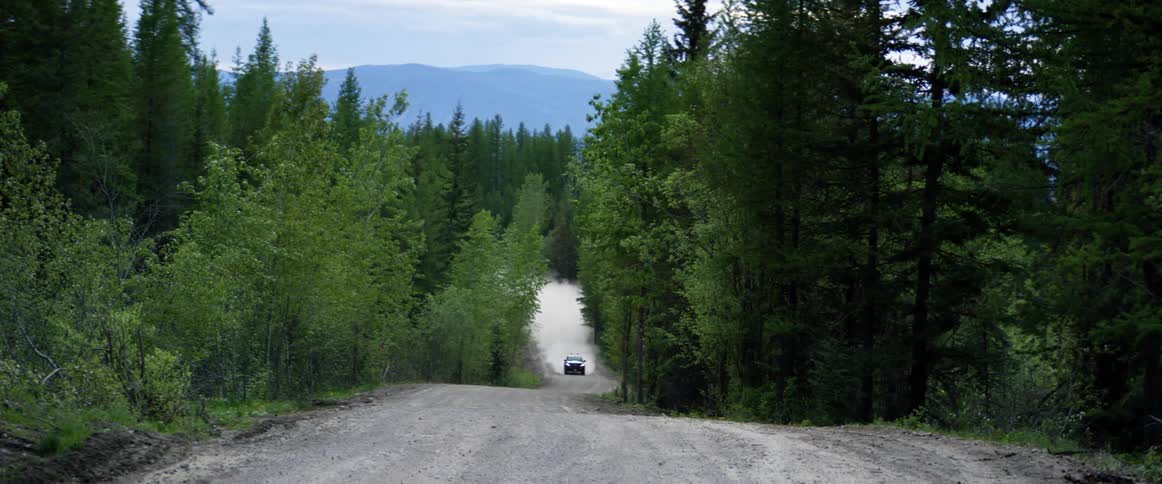 A vehicle kicks up dust as it drives down a narrow dirt road surrounded by forest