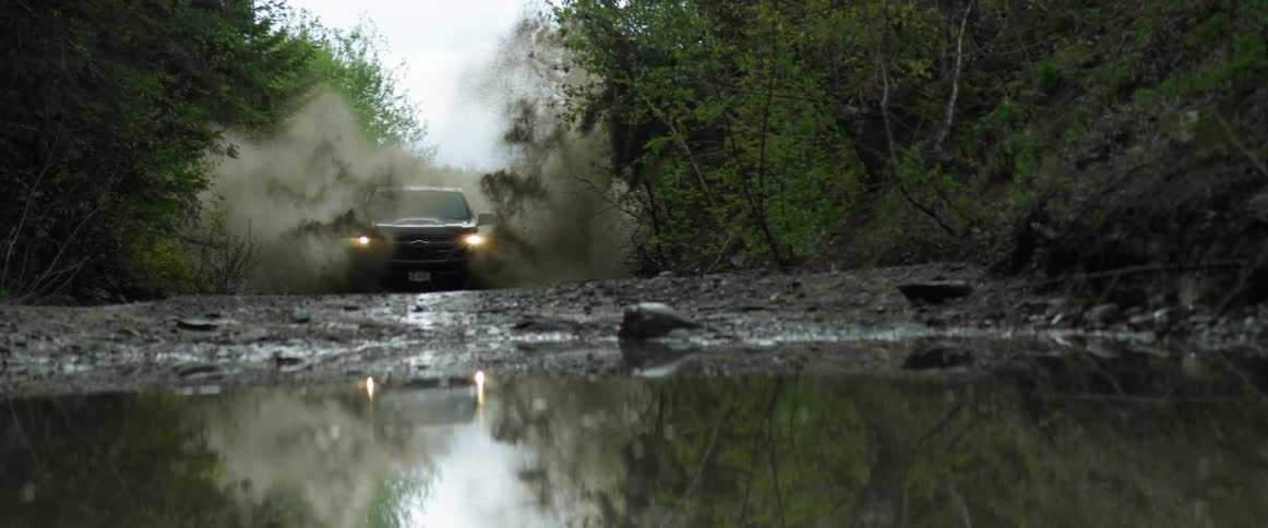 A truck drives through a large puddle in a heavily wooded rural area