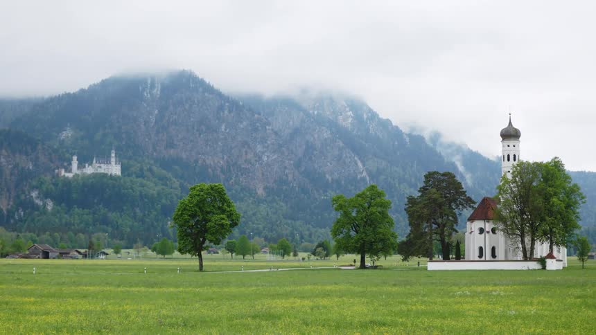 Baroque church of saint coloman in a field of green grass in front of mountains