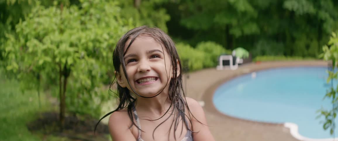 A little girl stands next to a swimming pool and smiles