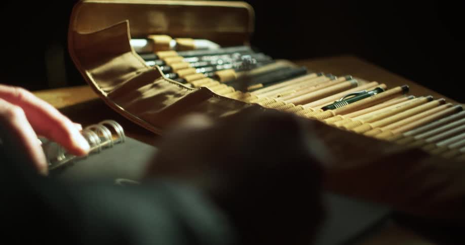 Someone draws with a pencil in a coiled notebook while a collection of pencils sits nearby