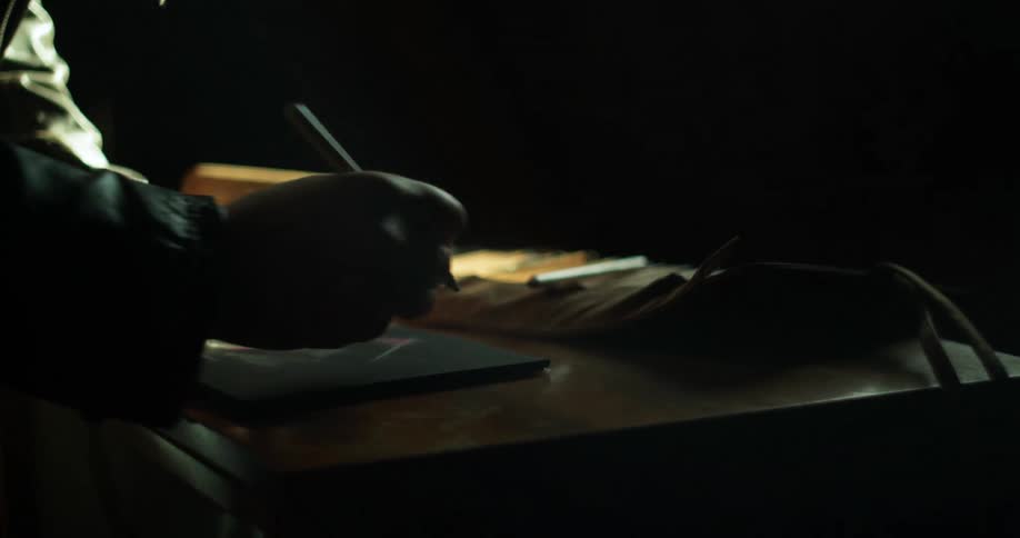 A man wearing a leather coat and gloves draws on a pad of paper in a dark room