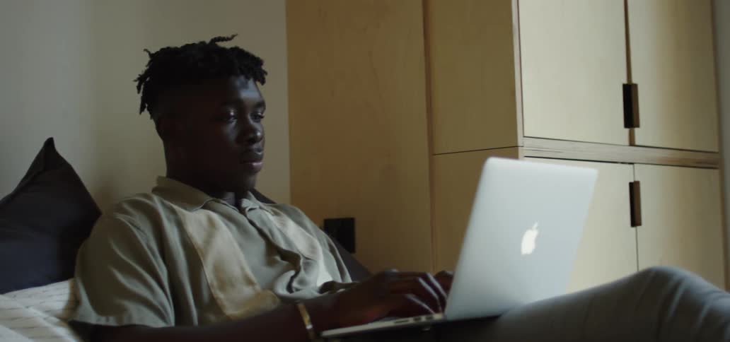 A black man sits in his bed and types on his laptop