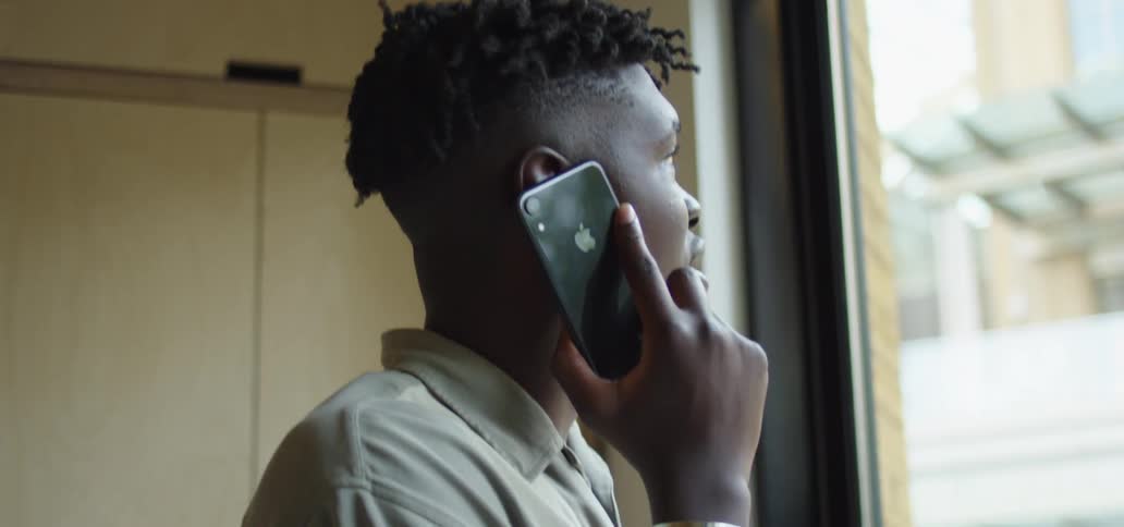 A man stands beside a large window looking outside on his cell phone