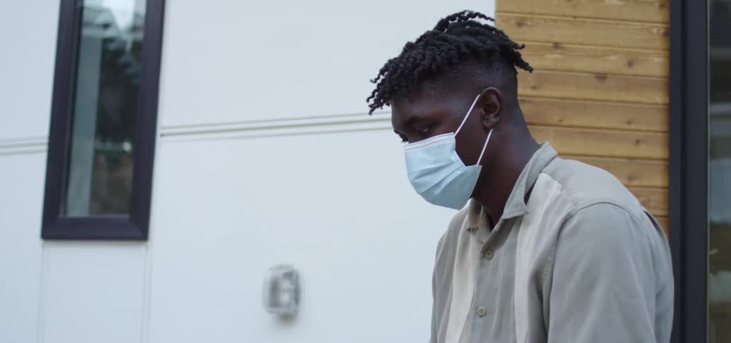 A teenager wearing a mask sits outside his house and uses a laptop