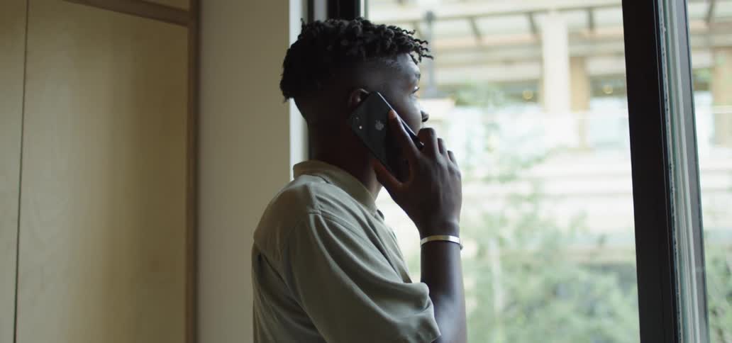 A black man stands indoors by a window and talks on a smartphone