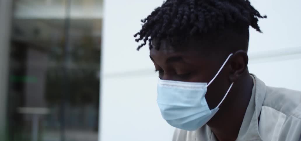 A teenager in a mask works on his laptop outside and stops to look around