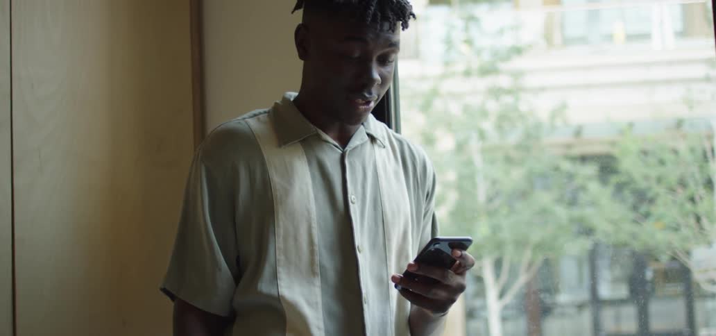 Teenage male standing next to a window is holding a smart phone and smiling while video chatting