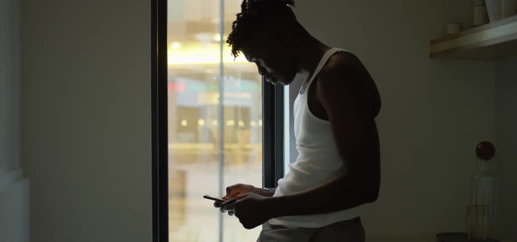 A man leaning against a counter near a long window looks down at his phone as he scrolls and types