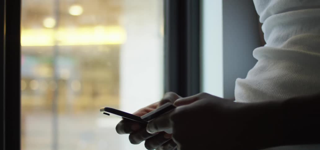 Male leaning against the wall next to a window holds a credit card while using his thumb to type on his smart phone