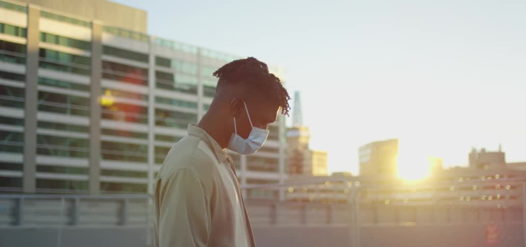 Young black man wearing face mask uses his cell phone while standing in the city at sunset