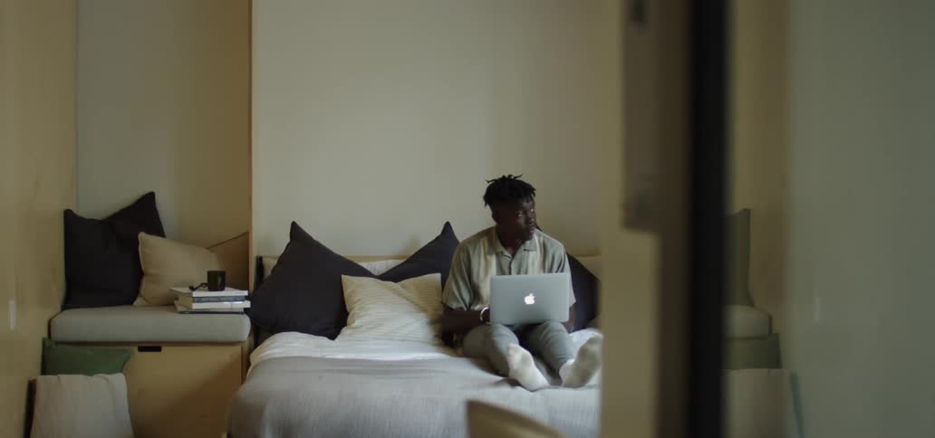Black man with grey polo shirt and pants sits on a bed and uses a laptop in the bedroom