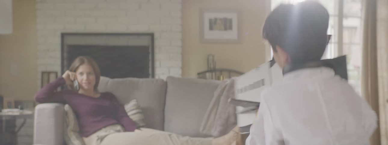 Mother sits on the couch while talking on the phone watching his son play with a toy in the living room