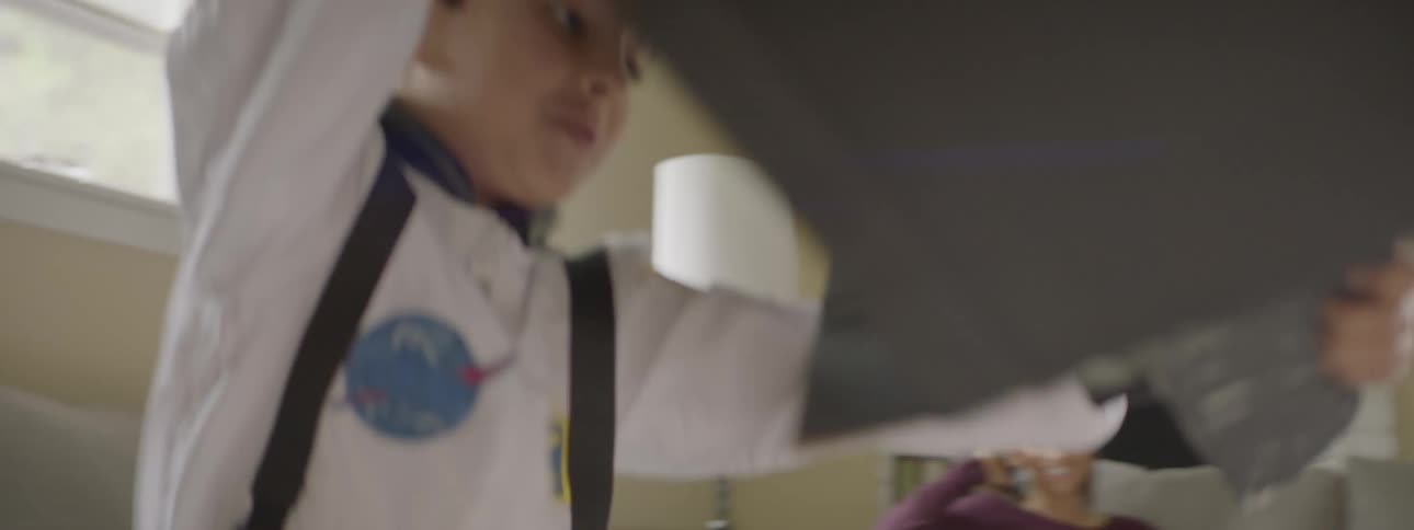 A young boy dressed as an astronaut runs around a living room with a box made into a space shuttle while his mother smiles and watches from a couch