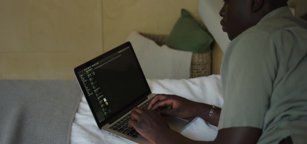 A teenage male is lying on his side on the bed while typing on a laptop